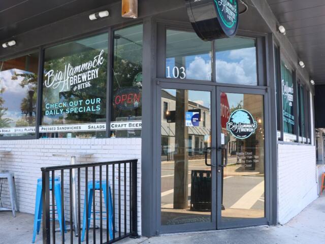 The exterior of a restaurant features signs reading, "Big Hammock Brewery & Bites." Outdoor seating lines the walls on a sidewalk with a small, black fence next to the entrance.