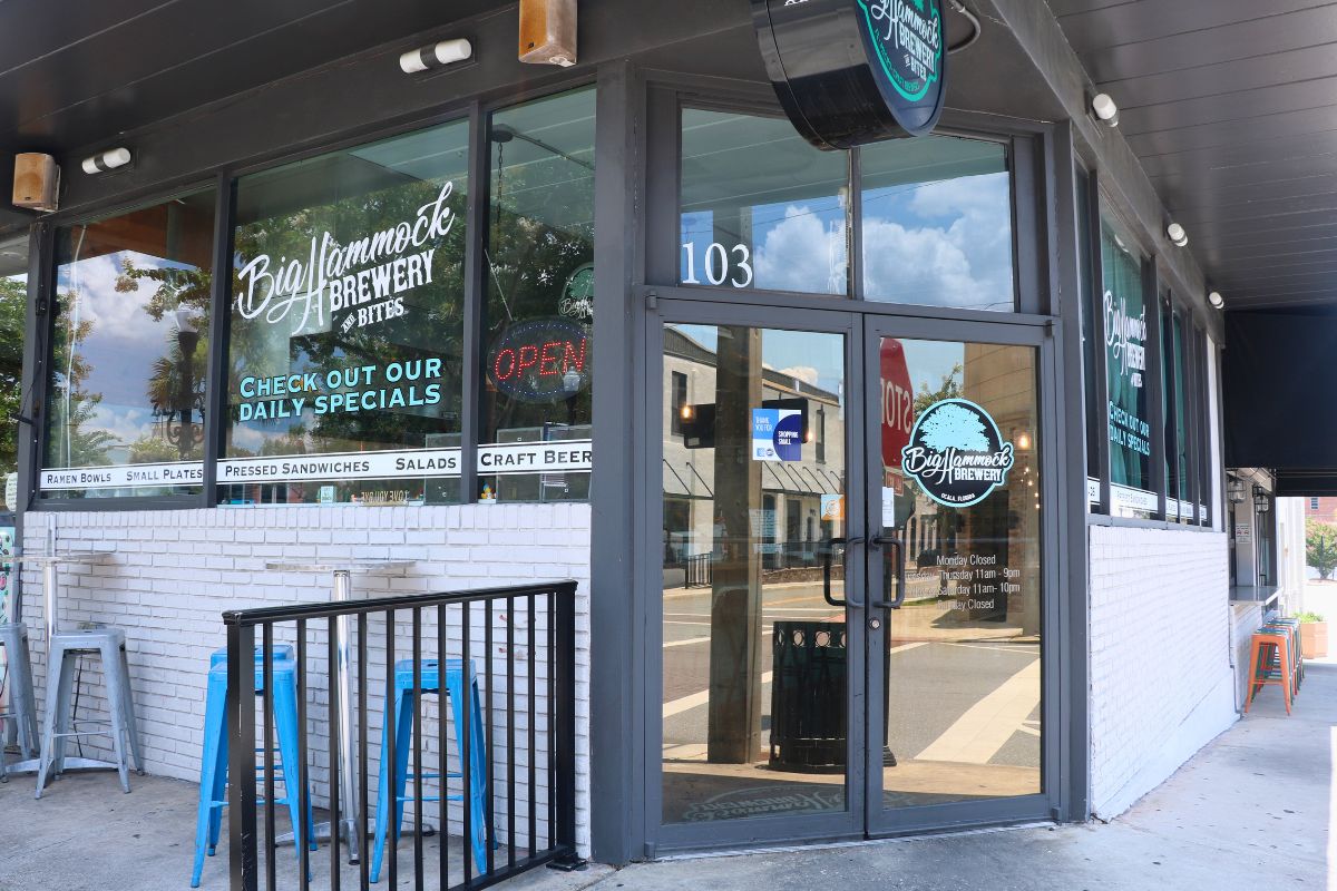 The exterior of a restaurant features signs reading, "Big Hammock Brewery & Bites." Outdoor seating lines the walls on a sidewalk with a small, black fence next to the entrance.