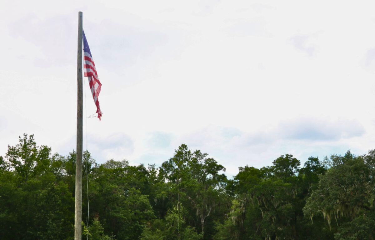 U.S. flag slightly waves in the wind on a flagpole in front of a tree line and blue, clowdy sky.