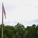 U.S. flag slightly waves in the wind on a flagpole in front of a tree line and blue, clowdy sky.