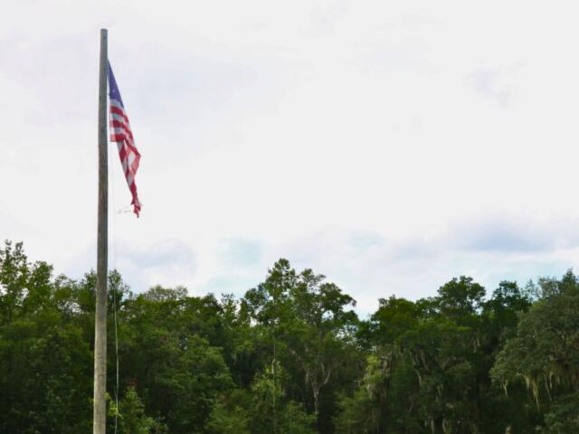 U.S. flag slightly waves in the wind on a flagpole in front of a tree line and blue, clowdy sky.