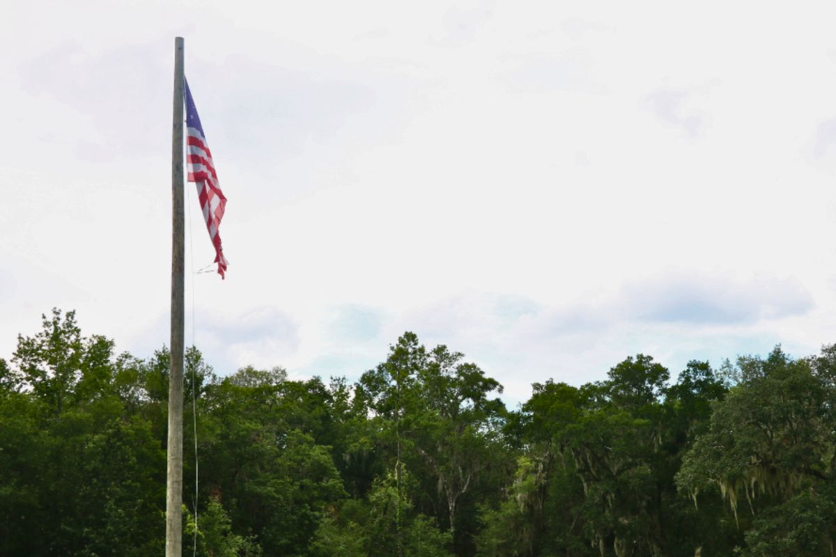U.S. flag slightly waves in the wind on a flagpole in front of a tree line and blue, clowdy sky.