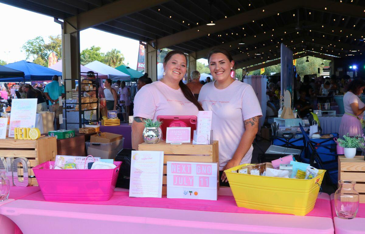 Two smiling women stand behind a pink-sashed booth at an outdoor market, with raffle baskets.