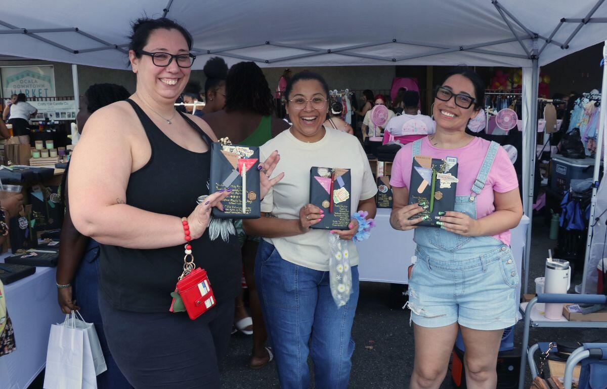 Three smiling women stand under a white canopy at an outdoor market, each holding a decorated black book.