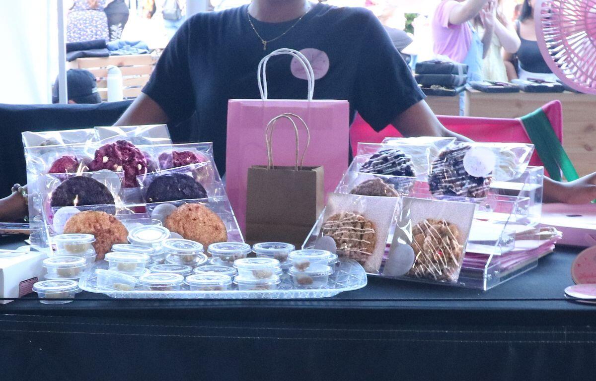 Vendor at a market stall displaying assorted cookies in clear acrylic cases with small sample cups on a black table, pink gift bags in the background.