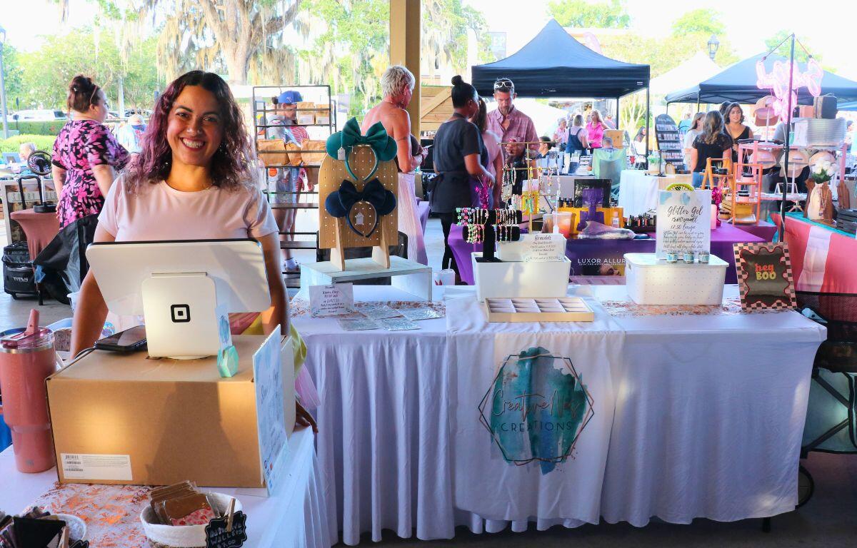 Smiling woman stands behind a checkout counter at an outdoor market stall selling jewelry and crafts.