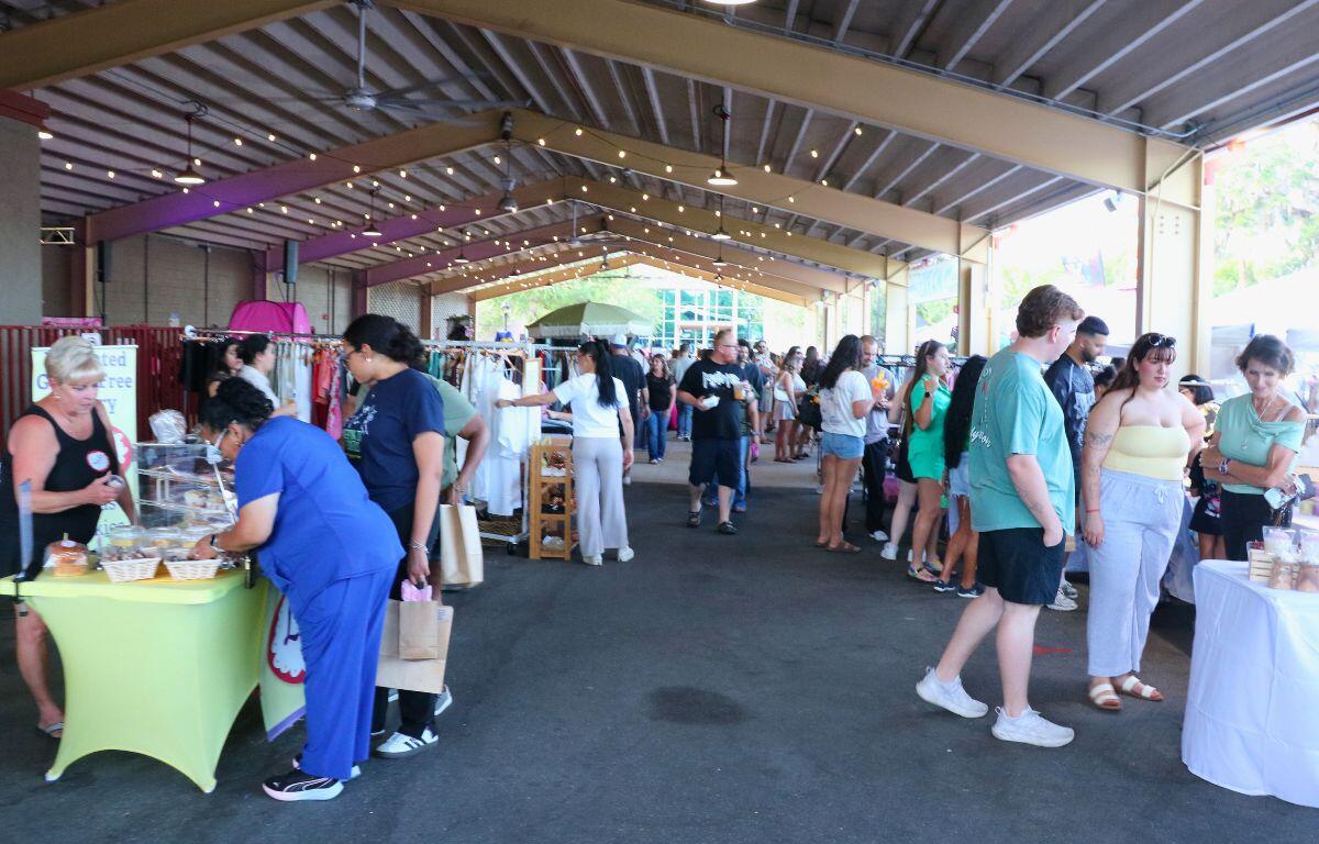 A crowd of people walk through an outdoor market under a large pavilion.