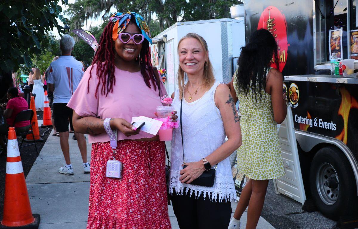 Two smiling women at a street festival; one in pink top and patterned red skirt with heart-shaped glasses, holding a pink drink.