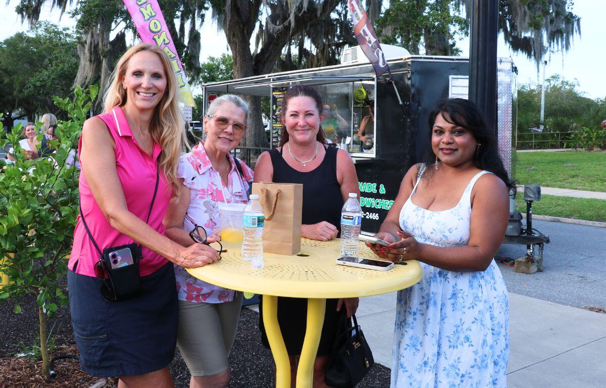 Four women stand around a yellow round table at an outdoor event, smiling near a food truck trailer and greenery behind them.