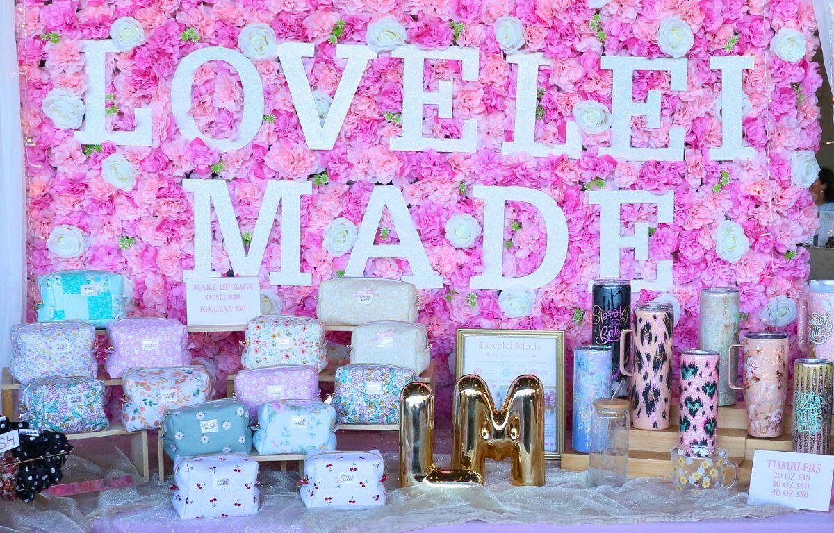 Pink flower-wall display with large white letters forming a sign; in front are pastel makeup bags, dressing pouches, and decorative tumblers, plus a gold LM decoration.