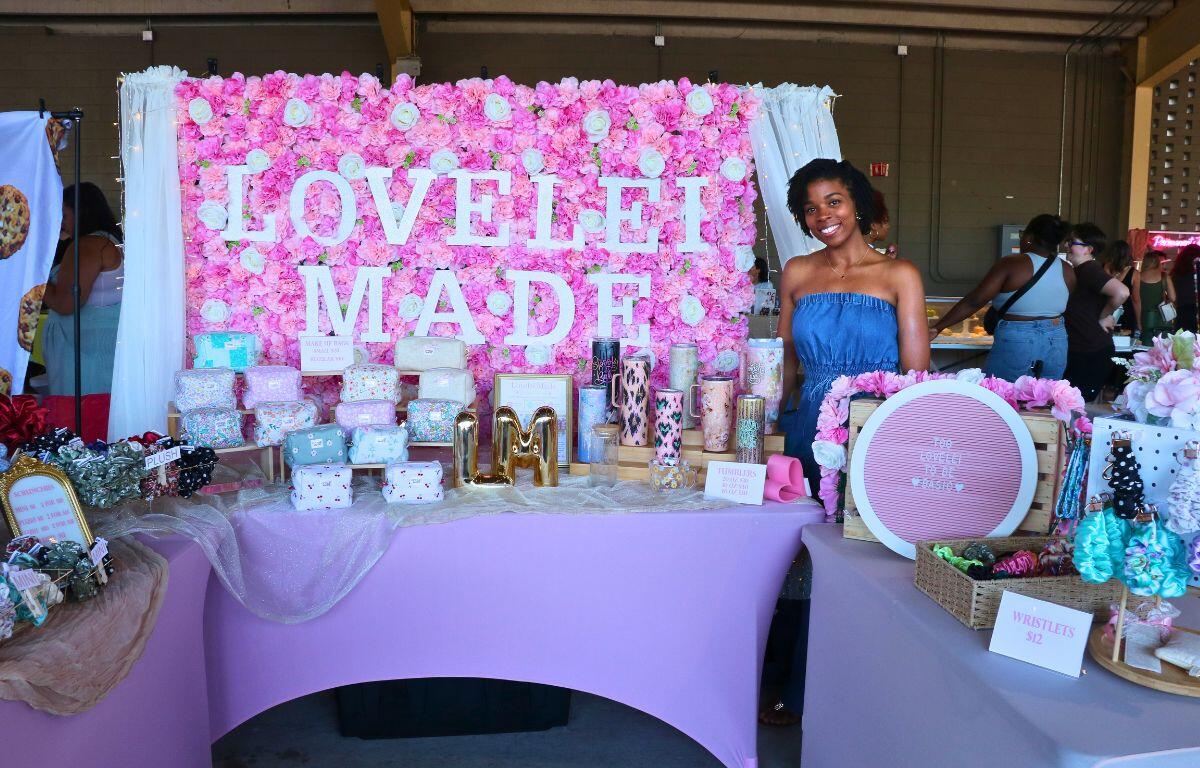 Vendor at a pink floral backdrop booth with a 'LOVE I MADE' sign, displaying wrapped gifts and accessories on a lavender table.