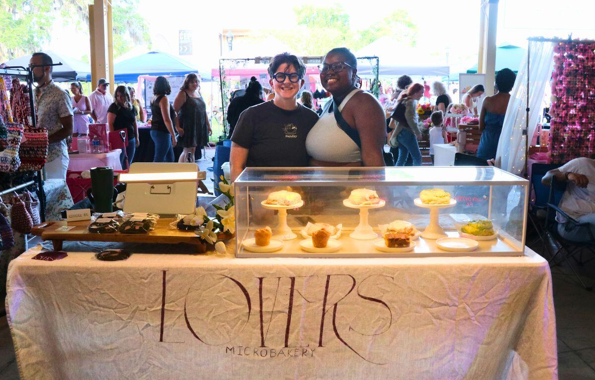 Two people stand behind a glass display case of cupcakes at a market stall, smiling at the camera with a cropped handwritten banner reading 'LOVERS Microbakery.'
