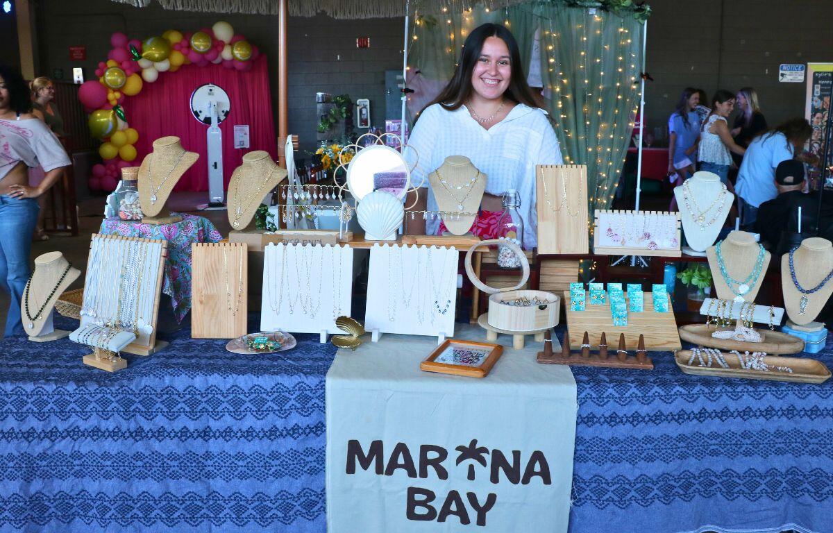 Jewelry vendor at a market stall with necklaces and bracelets on display stands; smiling woman behind the table.