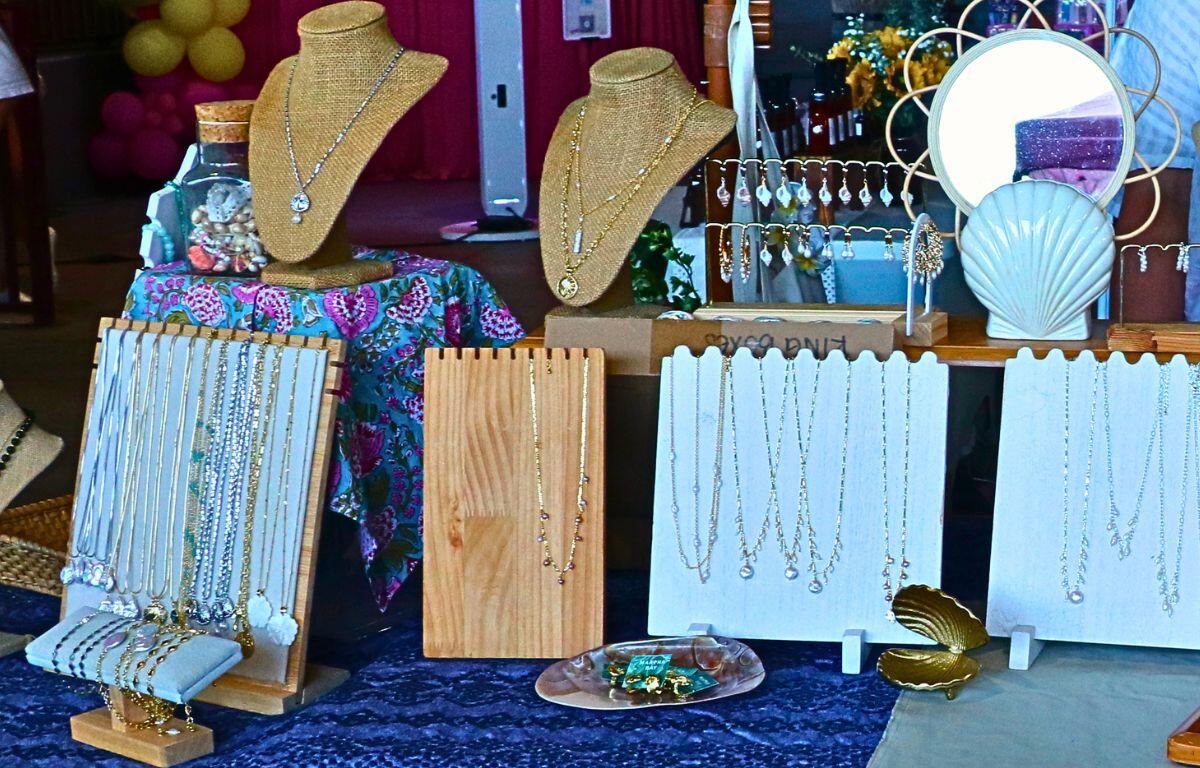 Jewelry display at a market stall: gold necklaces on bust stands, multiple chains and earrings on white boards, bracelets laid out, with shells and a patterned cloth backdrop.