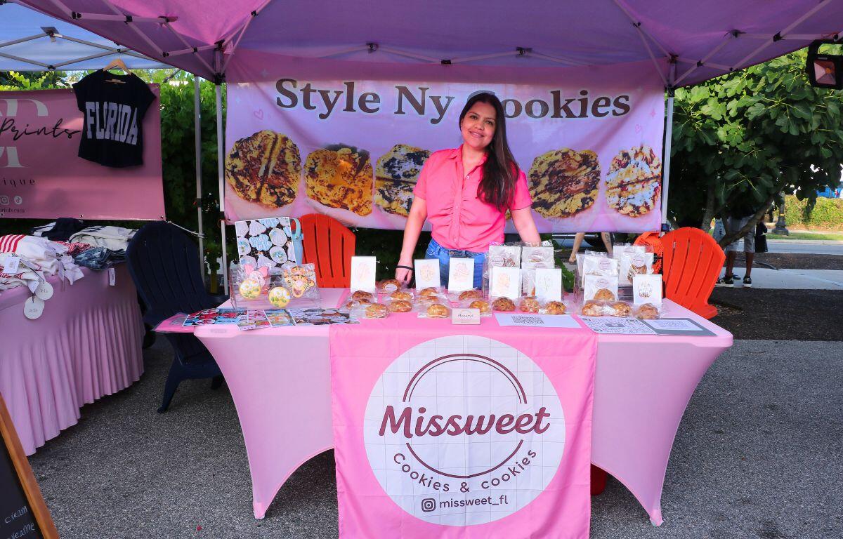 Woman seller stands behind a pink cookie booth labeled 'Misssweet Cookies & Cookies' with cookies on display under a pink tent.
