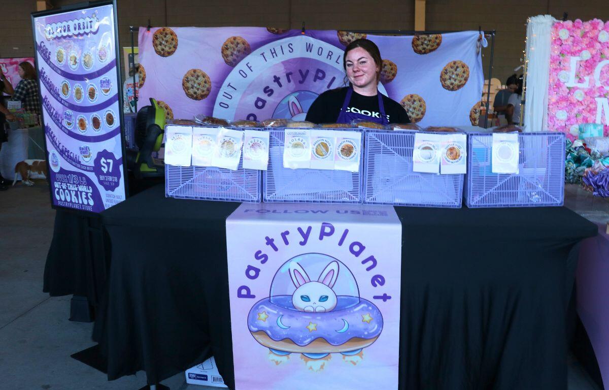 Vendor at a pastry booth with a purple donut banner and cookie display behind the table; logo reads PastryPlanet with a bunny in a donut design.