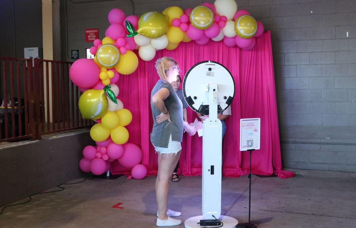 Woman in a gray top and white shorts uses a circular photo booth on a tall stand, with a bright pink balloon arch and pink backdrop.