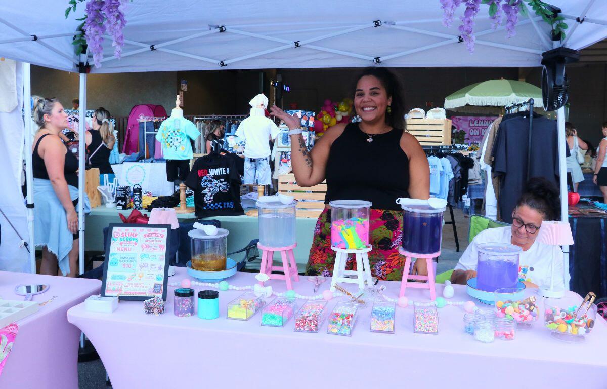 Smiling vendor standing behind a pastel pink candy bar at an outdoor market, with three drink dispensers and colorful toppings on the table.