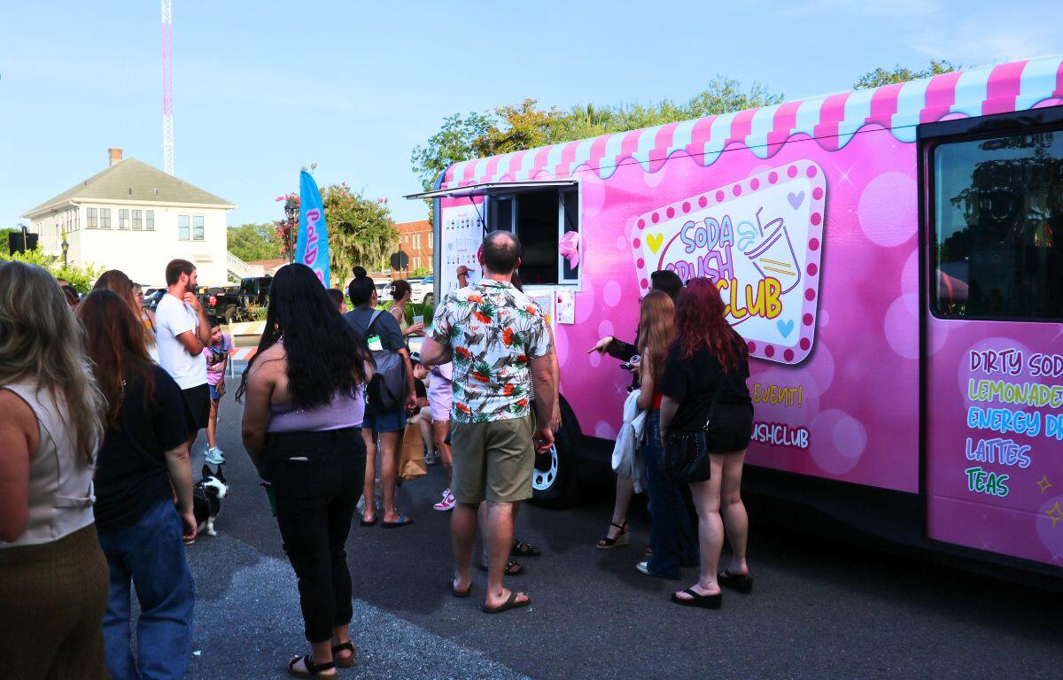 Bright pink soda truck with a striped canopy, a line of people waiting to order at an outdoor event.