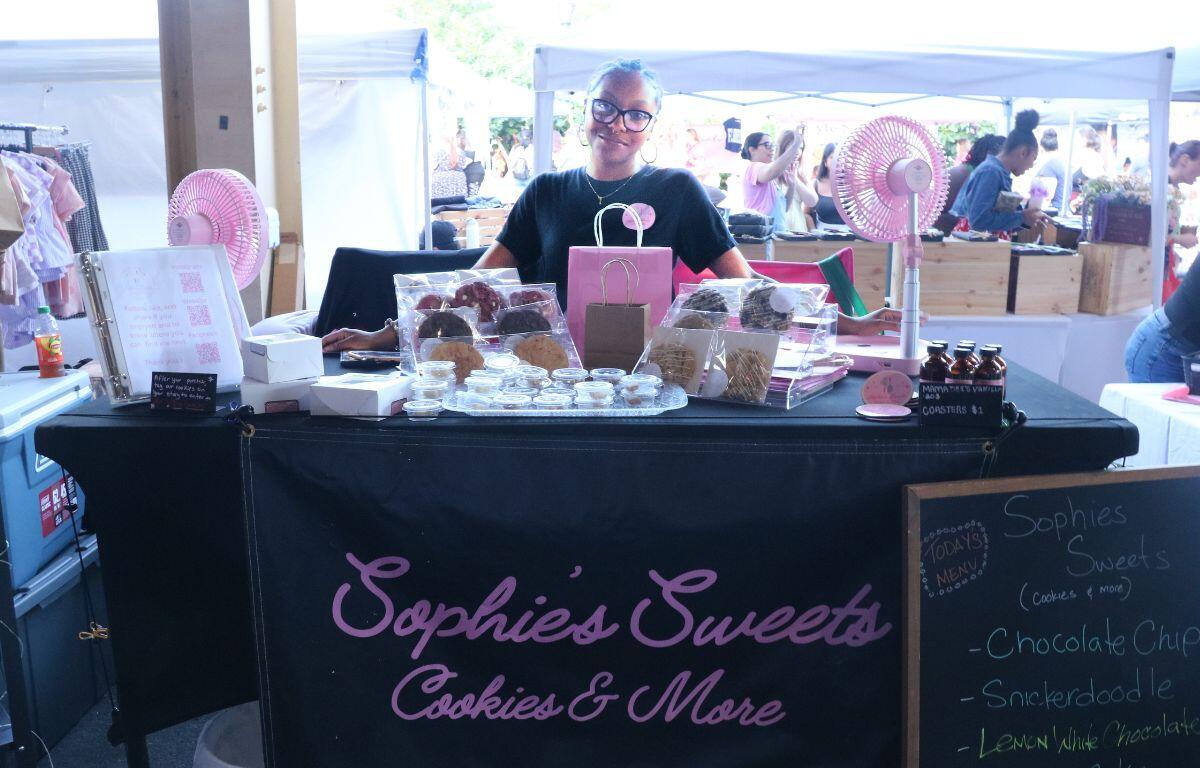 Vendor at a market stall named Sophie's Sweets Cookies & More, smiling behind a table of cookies and treats in clear bags.