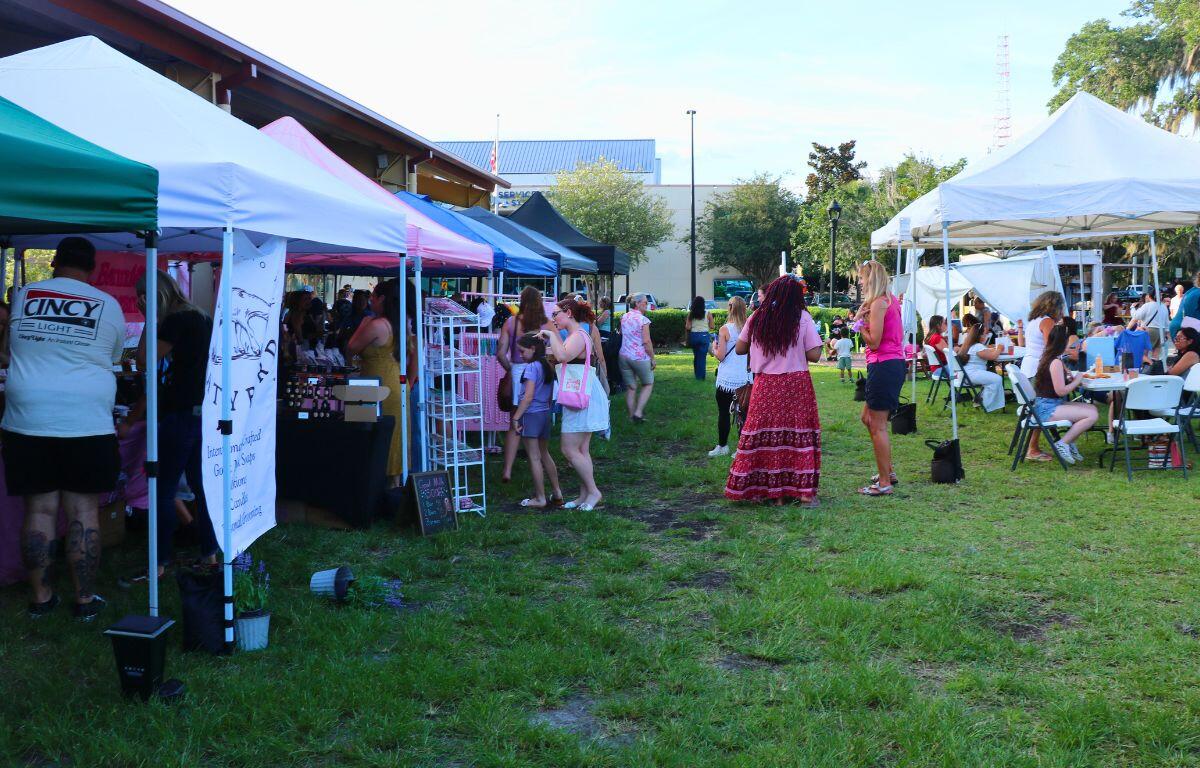 Outdoor market with colorful vendor tents and people browsing stalls on a grassy lawn.