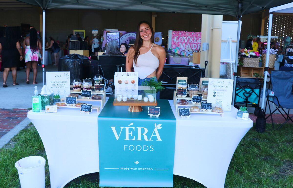 Smiling woman stands behind a teal bannered market stall that reads 'VÉRA FOODS,' with samples and cups on the table beneath a teal canopy.