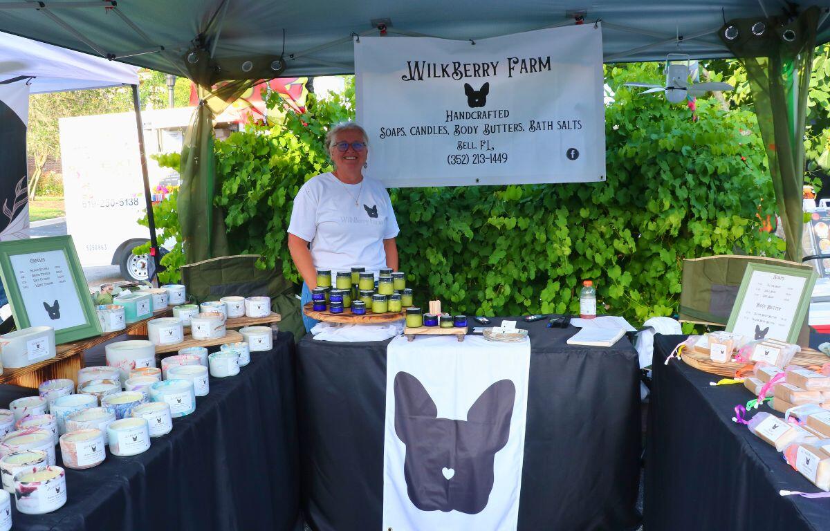 Vendor at WilkBerry Farm booth selling handcrafted soaps and body treats under a canopy, smiling.