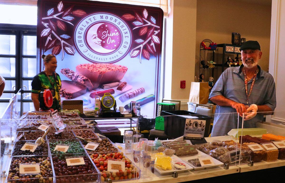 Chocolate confections are displayed in clear containers with two people behind the display.
