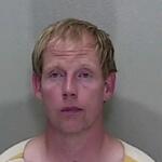 A mugshot of a man with short blonde hair in an orange and white striped jumpsuit.