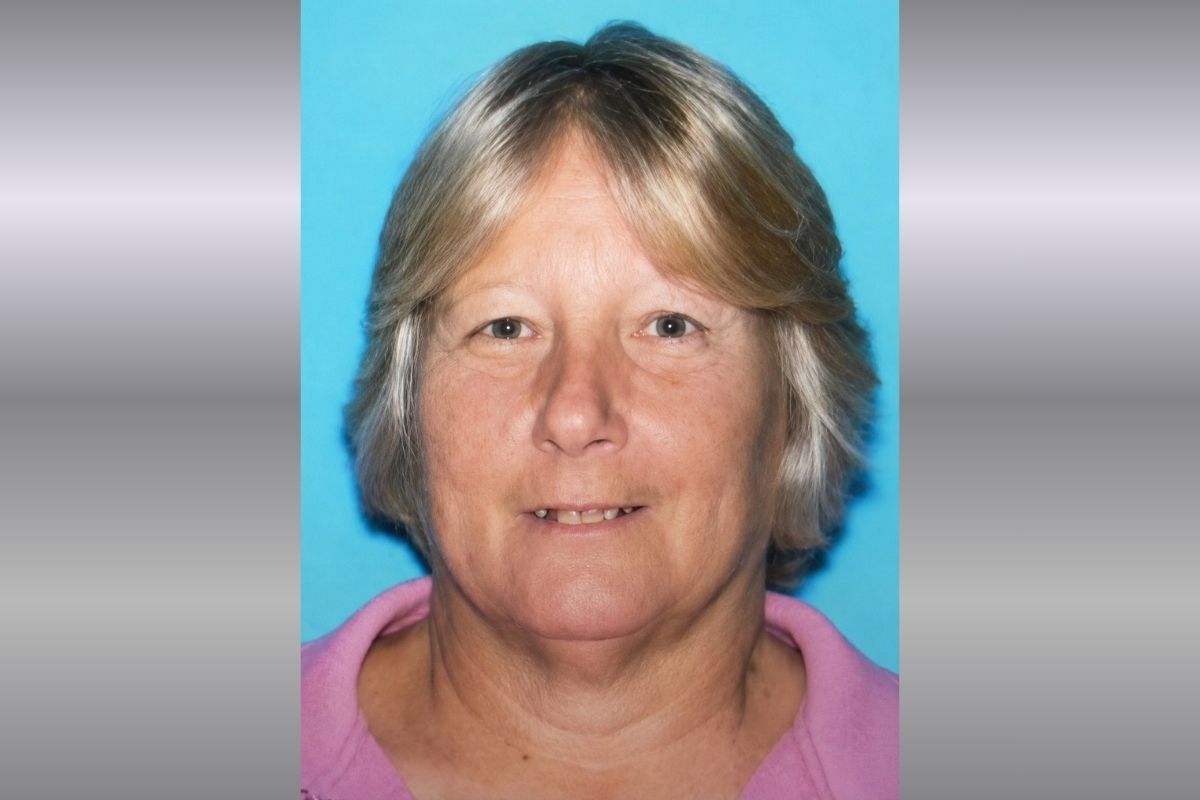 A woman with blonde-gray hair and a pink shirt against a blue and gray background.