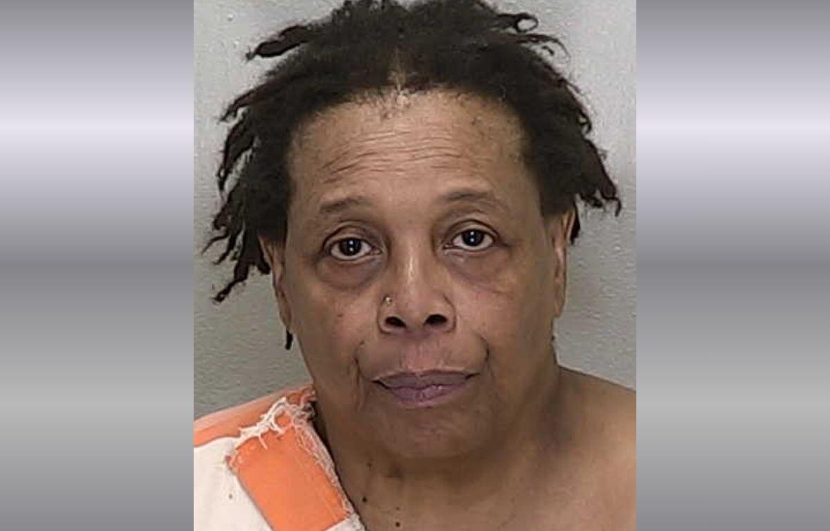 A mugshot of a black woman with short hair in an orange and white striped jumpsuit against a gray background.