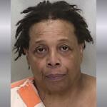 A mugshot of a black woman with short hair in an orange and white striped jumpsuit against a gray background.