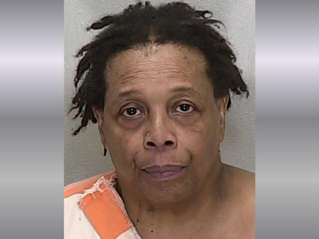 A mugshot of a black woman with short hair in an orange and white striped jumpsuit against a gray background.