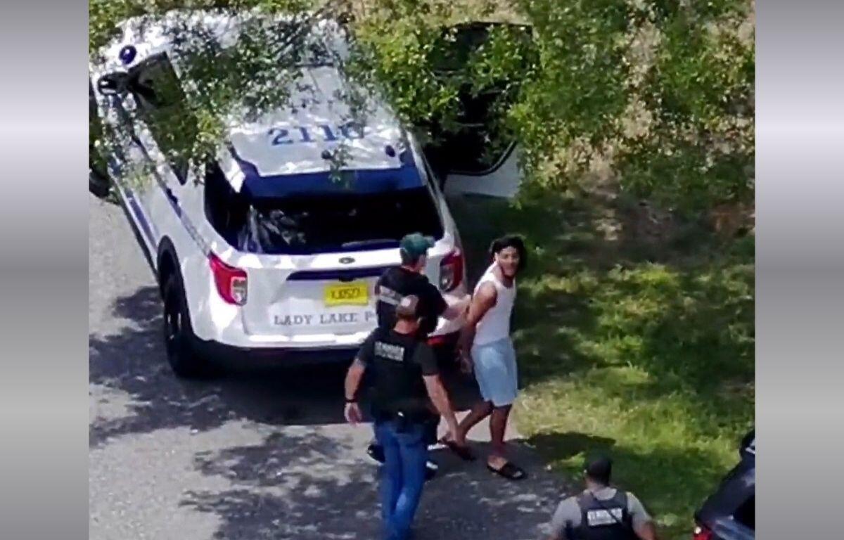 A still for drone surveillance footage of a man in a white tank top and blue shorts being arrested by law enforcement officers.