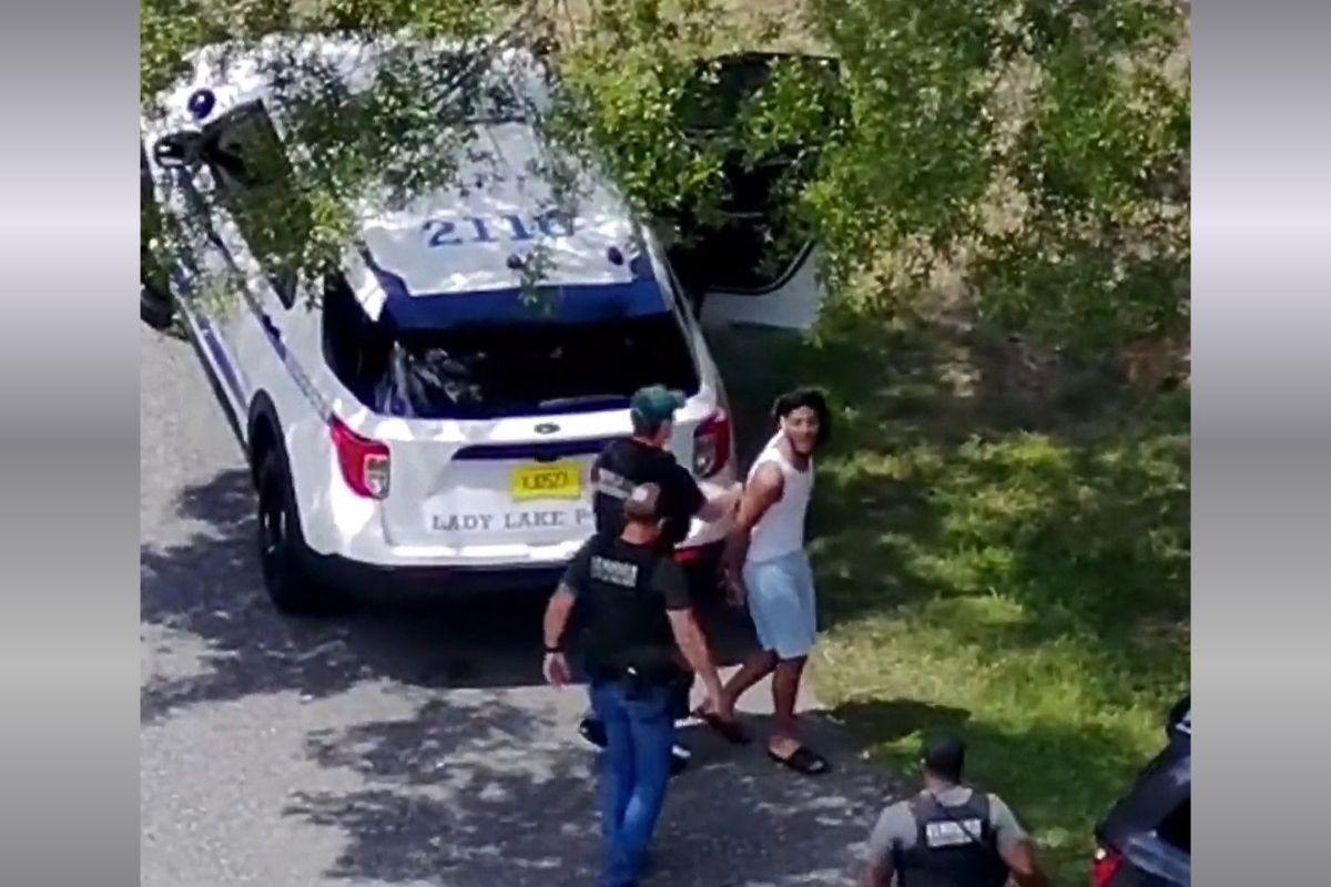 A still for drone surveillance footage of a man in a white tank top and blue shorts being arrested by law enforcement officers.
