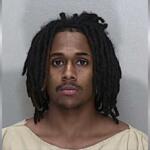 A mugshot of a African American man with shoulder-length black dreadlocks in a beige jumpsuit.