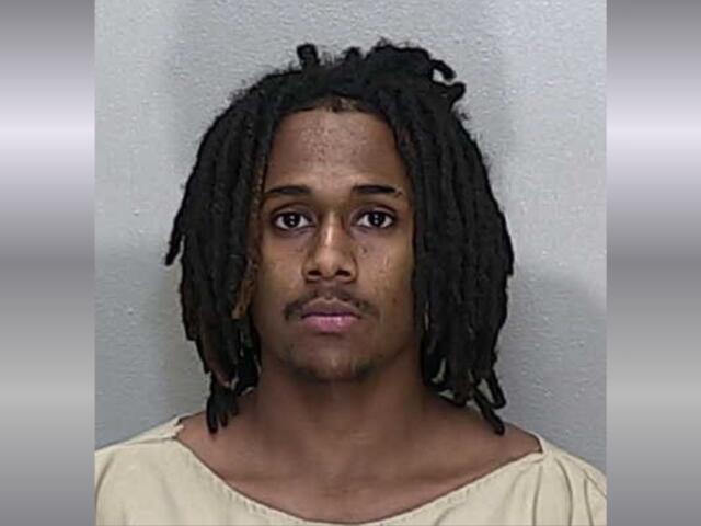 A mugshot of a African American man with shoulder-length black dreadlocks in a beige jumpsuit.
