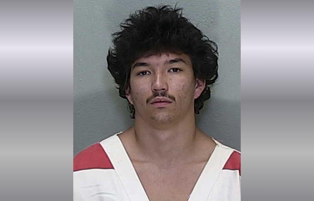 Mugshot of a young man with dark, tousled hair and a thin mustache, wearing a white and red striped jail uniform against a gray backdrop.
