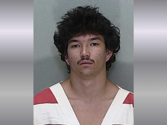 Mugshot of a young man with dark, tousled hair and a thin mustache, wearing a white and red striped jail uniform against a gray backdrop.