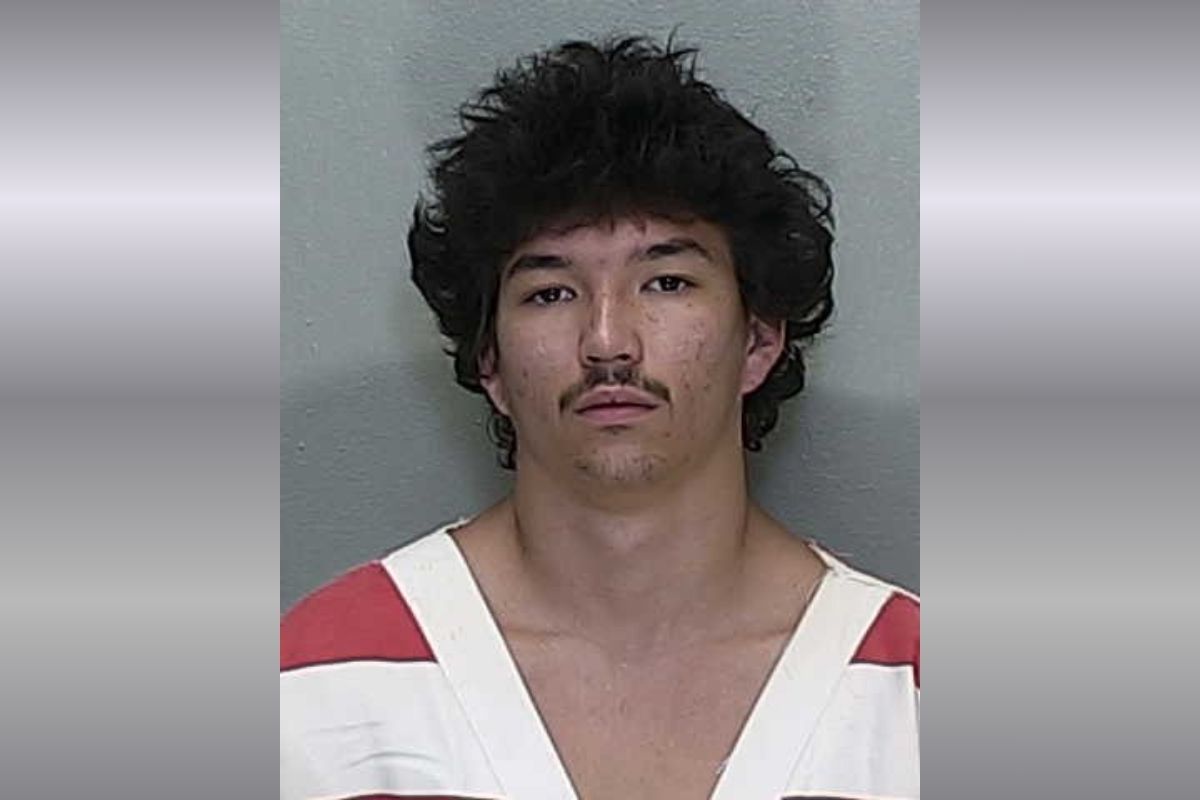 Mugshot of a young man with dark, tousled hair and a thin mustache, wearing a white and red striped jail uniform against a gray backdrop.
