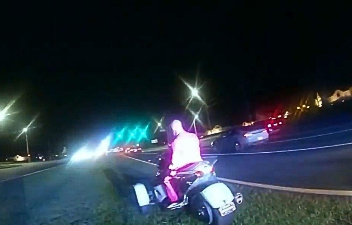A still from body cam footage of a man in a pink business suit sitting on a silver trike.