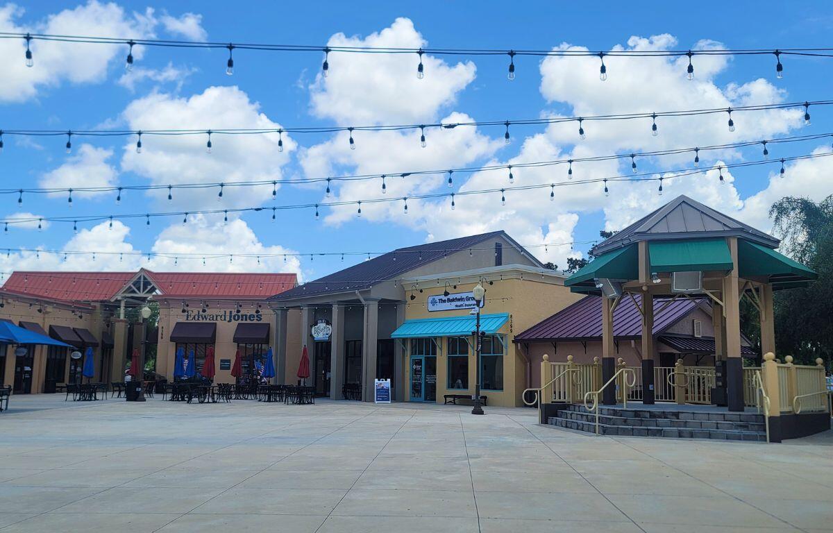 Open-air shopping plaza with string lights, storefronts (including Edward Jones), outdoor seating under umbrellas, and a gazebo against a blue sky.