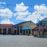 Open-air shopping plaza with string lights, storefronts (including Edward Jones), outdoor seating under umbrellas, and a gazebo against a blue sky.