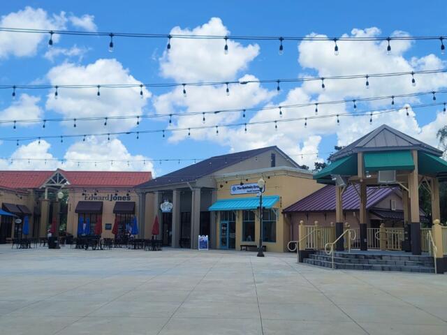 Open-air shopping plaza with string lights, storefronts (including Edward Jones), outdoor seating under umbrellas, and a gazebo against a blue sky.