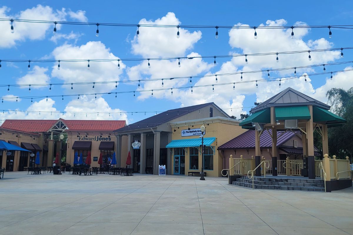 Open-air shopping plaza with string lights, storefronts (including Edward Jones), outdoor seating under umbrellas, and a gazebo against a blue sky.