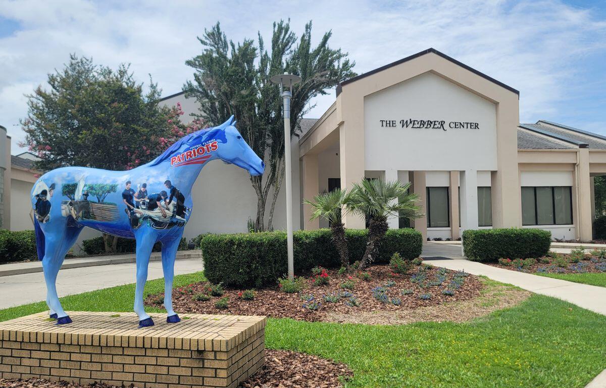 A large horse statue with painted depictions of medical students stands in front of a large conference center. Over the entrance of the center reads, "The Webber Center."