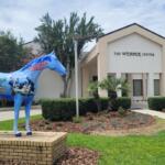 A large horse statue with painted depictions of medical students stands in front of a large conference center. Over the entrance of the center reads, "The Webber Center."