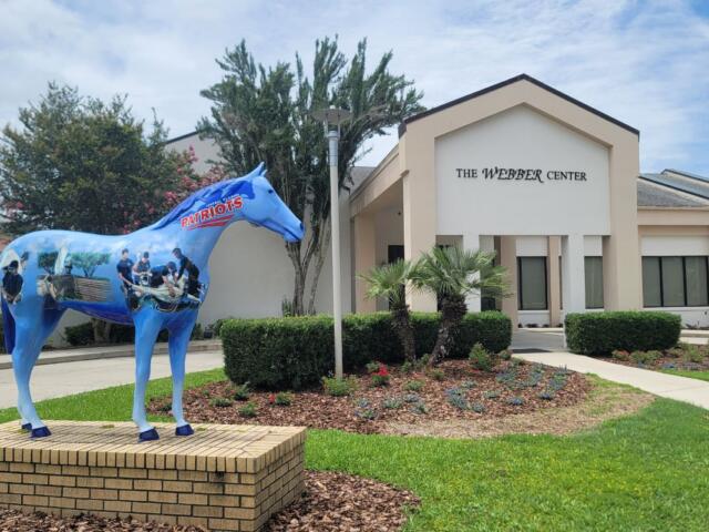 A large horse statue with painted depictions of medical students stands in front of a large conference center. Over the entrance of the center reads, "The Webber Center."