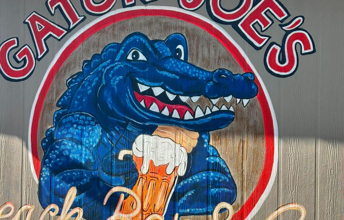 Sign for Gator Joe's with a blue alligator holding a beer inside a circular red-and-white logo, blue sky above.