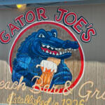 Sign for Gator Joe's with a blue alligator holding a beer inside a circular red-and-white logo, blue sky above.